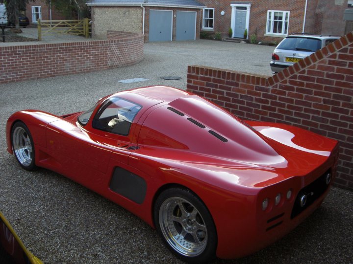 Garagenearly Pistonheads Months Finished - In the foreground of the image, a striking red sports car is parked. The car appears to be an electric model, judging by the unique aerodynamic body. Its sleek design and shiny finish reflect the light, suggesting a polished exterior. The vehicle is parked on a gravel area next to a brick wall, adding a rustic touch to the scene. 

In the background, there is a domestic building with a sturdy brick facade and a contrasting white door. Adjacent to it, a wooden fence with brick pillars runs along the street. A car and a truck are parked in the same vicinity as the sports car, blending into the everyday suburban backdrop.