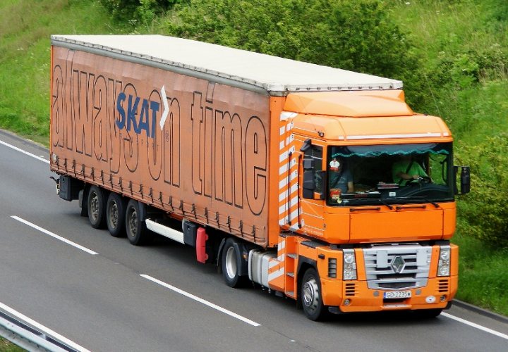 Made me laugh - Page 1 - Commercial Break - PistonHeads - The image features a semi-trailer truck in motion. The cab of the truck is painted a vibrant shade of orange, contrasting with its silver grill. The truck occupies most of the frame, traveling on what appears to be a two-lane highway with a grassy area and trees in the background. The truck is pulling a trailer, likely for transporting goods. The driver of the truck can be seen in the cab, and there is a logo on the side of the truck which reads "SKTRA."