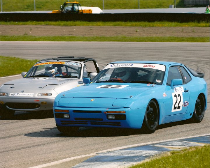 Porsches Pistonheads - The image captures an exciting moment on a winding race track where two race cars, one blue and the other silver, are driving closely together. Both cars have the number 13 visible on their hoods, indicating a possible competitive event or identification number. The setting also suggests a professional racing environment, with a crowd of spectators visible in the background, adding to the intensity and excitement of the scene. The race cars' aerodynamic design and gear suggest high speeds, further emphasizing the competitive nature of this deep focus photograph.