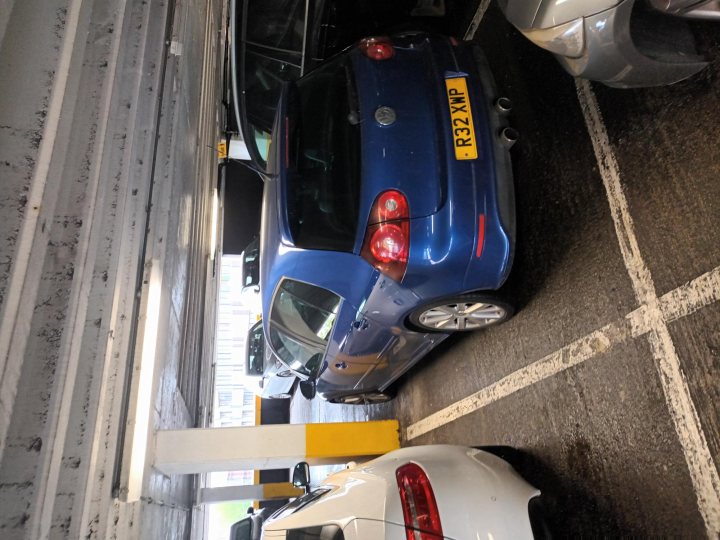 MK5 Golf R32 - number 3.. - Page 4 - Readers' Cars - PistonHeads UK - The image shows an indoor parking garage with multiple cars parked. A blue sedan is prominently featured in the foreground, parked adjacent to a white hatchback. To the left of the frame, there's a black vehicle, and on the right side, there's another blue car partially visible. The garage has a concrete floor and is lit by artificial lighting. The structure of the parking garage includes columns supporting the ceiling, which appears to be made of metal. There are no visible people or distinctive landmarks in the image.