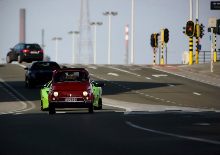 A car is stopped at a red light - Pistonheads - This image captures a dynamic road scene. It features two vintage cars in motion on a highway. The foreground car, a neon red convertible, is followed by another car, possibly neon green and yellow. The road is equipped with multiple traffic lights at various distances, indicating a regulated and controlled traffic system. The traffic suggests it's daytime with clear visibility. The overall scene is harmonious, with the cars moving smoothly and the traffic lights indicating an orderly flow of traffic. The presence of these vintage cars may imply a nostalgic or tourist-oriented environment.