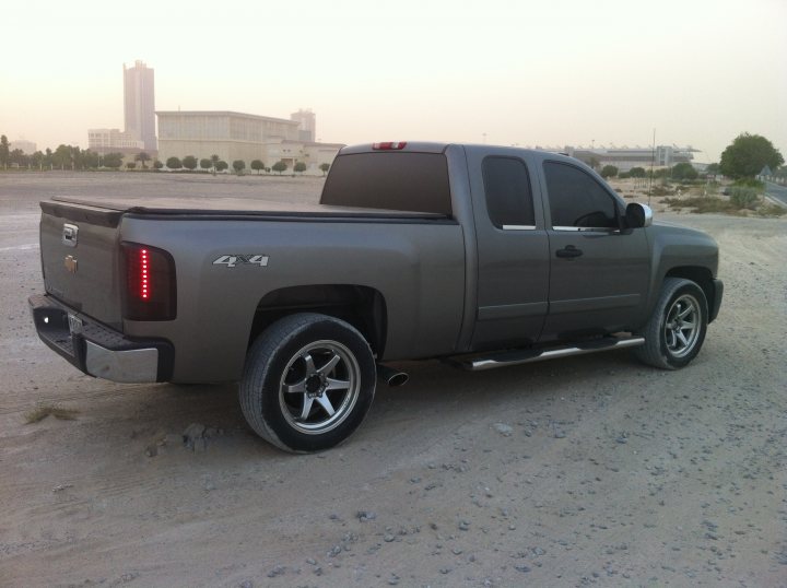 Not what  I had in mind but ....... - Page 1 - Yank Motors - PistonHeads - This image features a full-sized pickup truck parked on what appears to be a dirt surface. The truck is in profile view facing to the right, highlighting its robust build and features a gray color scheme. The vehicle has a prominent tinted-over back window and taillights that include a prominent red oval with a black border, possibly indicating a specific model or brand. The background shows a vast landscape with a city skyline, suggesting the truck is in a remote or rural area with a view to an urban setting in the distance.