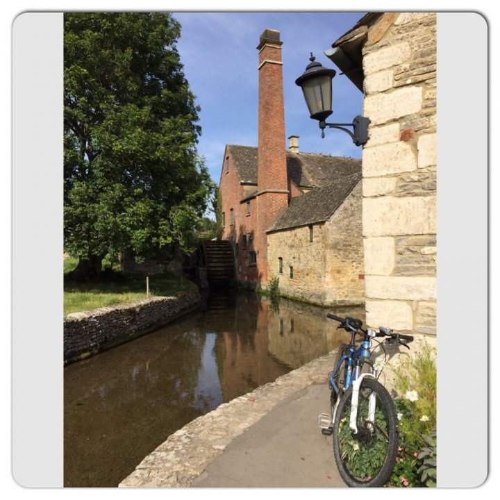 The "Photos From Today's Ride" thread... - Page 225 - Pedal Powered - PistonHeads - The image depicts a serene outdoor setting, featuring a brick building with a massive chimney and a stone exterior. A single lamp post is situated near the building, illuminating the area. A tranquil water feature can be spotted in front of the building, likely a part of a moat or small canal. Near the water, a blue bicycle is parked, leaning against a rail, adding a touch of everyday life to the scene.