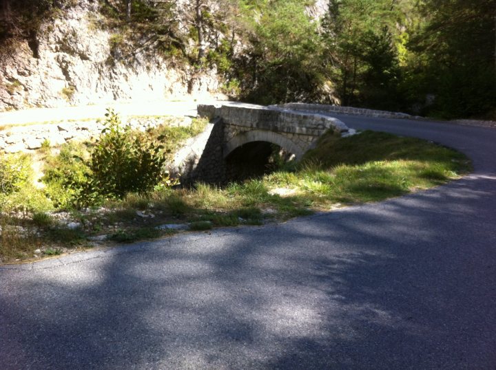 Driving France - Page 4 - Chimaera - PistonHeads - The image presents a serene outdoor scene. A sturdy stone bridge arching over a river or road cuts through a lush, grassy landscape. The bridge, constructed from layers of evenly placed stones, connects two sections of a winding dirt path which meanders through the grassy area. The path appears to be a quiet trail, used by only a few. In the distance, a rocky cliff face rises, contrasting with the flat slab of the smooth asphalt road. The surrounding environment suggests a peaceful rural setting, possibly in a region known for its natural beauty.