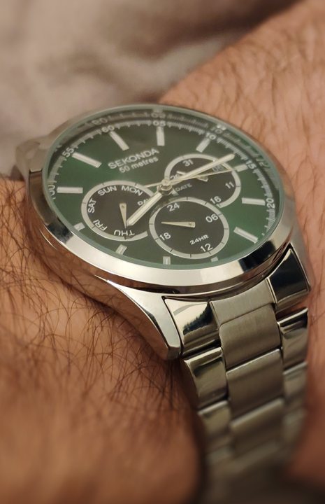 The image showcases a person's wrist with a watch that features two clock faces, one at the top and another at the bottom. The watch has a sleek design with a dark strap, a green bezel, and a silver case. It also displays the time '5:10' on both faces of the watch. The person is wearing a wristwatch with multiple complications or features such as dual time zones, chronographs, or moon phases.