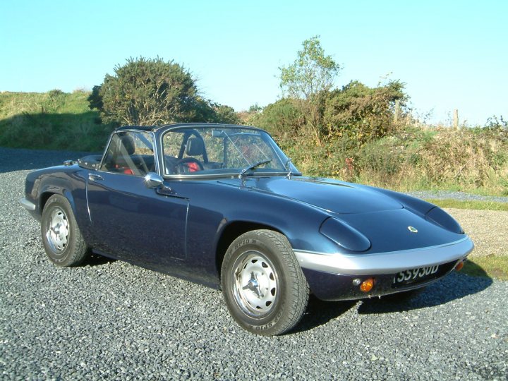 lotus servicing, South Wales - Page 1 - South Wales - PistonHeads - The image is a photograph of an older blue convertible sports car parked on a gravel surface with sparse vegetation in the background. The car has a unique, aerodynamic body style with a removable top and long, narrow headlamps. Its side mirrors and door handles are visible, and there's a distinctive air intake behind the front wheel. The car features round silver hubcaps and two exhaust pipes at the rear. The gravel ground shows signs of use, and the weather appears to be sunny. There are no visible texts or distinctive markings on the car.