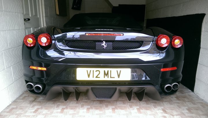 I have joined the club - Page 1 - Ferrari V8 - PistonHeads - The image shows a sleek two-door car parked inside a garage. The car is black with a distinctive hood that has two vents. The license plate reads "V12 MLV". The car is equipped with red taillights and side mirrors. The garage features a white wall and part of an open door can be seen in the background.