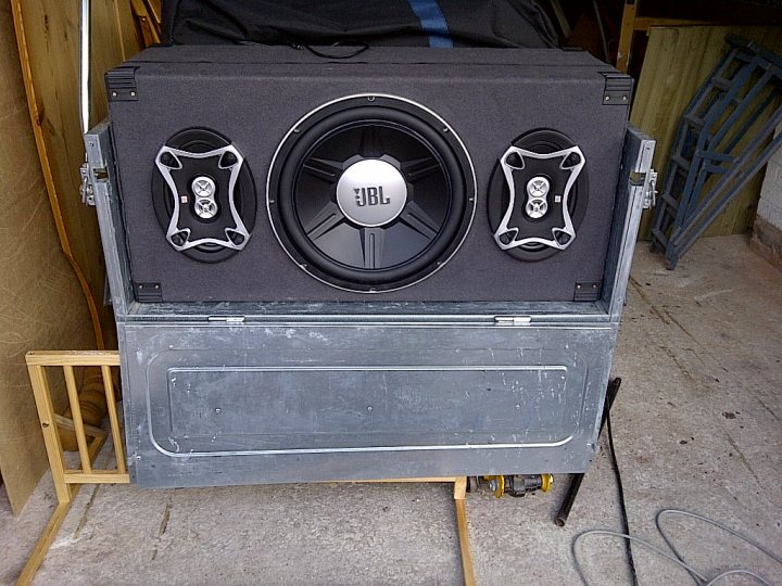 Music in a field - Page 1 - Tents, Caravans & Motorhomes - PistonHeads - The image showcases a large gray workspace lying on its side. It dominates the scene, with its right end visible. Two round 12-inch black speakers are attached to the workspace. Each speaker has a braided cable and a silver knob, reflecting the models brand. The object is set against the backdrop of a wooden wall, adding to the industrial aesthetic. The workspace is also equipped with wheels, suggesting its mobility.