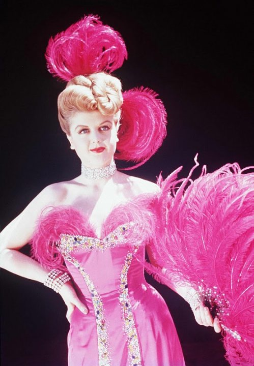 In this vintage style photo, a woman is the focal point, appearing confident and stylish. She is adorned in an elaborate pink dress embellished with flamingo-like feathers that match her pink feathered top hat. Her neckline is elegantly covered with a crystal beaded necklace, adding a touch of sophistication to her ensemble. Her pose and expression exude a sense of nostalgic glamour.