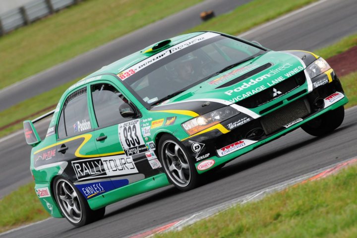 Here we go again.... Targa 2009 - Page 2 - New Zealand - PistonHeads - The image is a high-resolution, outdoor photograph with a detailed focus on a green car on a track. The car has an abundance of stickers and sponsorship decals visible on its body, indicative of a racing or rally car context. The side of the door showcases flags representing various crews or teams, suggesting competition at a professional level. The ground is made of asphalt, worn with tracks and curves, typical of a vehicle racing course. Surrounding the track, there is grass and a line of trees in the background.