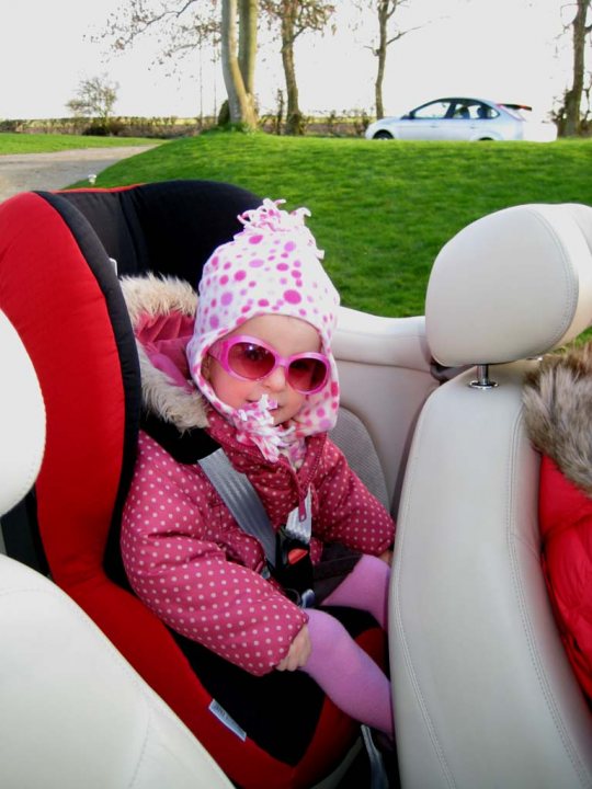 The image captures a delightful moment of a child sitting in the back seat of a car. The child is bundled up in a pink jacket and hat, which contrast with the car's beige interior. A pink and white polka dot hat is also worn by the child, adding a touch of playfulness. The car is parked next to a grassy field under a clear blue sky, suggesting an outdoor setting. The scene conveys a sense of warmth and innocence, likely on a day filled with excitement and adventure for the little one.