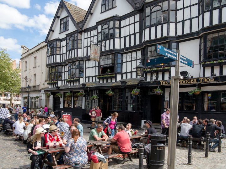 A group of people sitting on a bench in front of a building - Pistonheads - The image captures a lively scene on a city street in what appears to be a historical neighborhood. A large pub or tavern with a black and white facade dominates the view, complete with wooden beams and a tower-like structure on the upper right. The pub is bustling with people sitting at plates, enjoying their meals and drinks. A blue sign hanging from the building displays the name "Lindau Tram", possibly indicating nearby transportation or street name. The sky above is a clear blue, suggesting good weather, and the street is lined with a variety of other shops and buildings, contributing to the urban atmosphere.