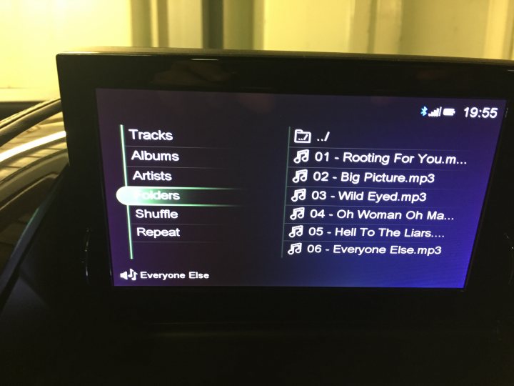 A flat screen tv sitting on top of a wooden table - The image presents a close-up view of an electronic device, which appears to be an old mp3 player. The interface is displayed on the screen, showing a list of songs on shuffle repeat play mode. The time stamp in the top right corner reads "19:55", suggesting that the photo was taken at that time. The current selected song is "Everyone Else...", indicating some form of classic rock theme or playlist. The background is dark, further emphasizing the device and its bright screen.