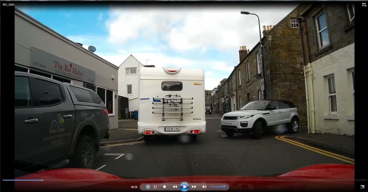 Dashcam on Boxster? - Page 1 - Boxster/Cayman - PistonHeads - The image depicts an urban scene featuring a white camper van in the middle of a narrow street. This is flanked on either side by parked cars and buildings, suggesting a dense cityscape. The street is lined with sidewalks and appears to have traffic calming measures in place. Above the road, there are clear blue skies and a few scattered clouds, indicating fair weather.