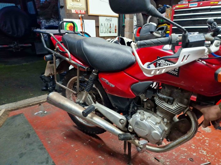 Riding around the world... on an inappropriate bike!  - Page 2 - Biker Banter - PistonHeads UK - The image shows a red motorcycle parked inside a garage or workshop. The motorcycle is the central focus of the photo, taking up most of the frame. It's positioned in front of what appears to be a workbench with various tools and parts scattered on it. The room has an industrial feel, with its concrete floor and utilitarian furnishings. The lighting is subdued, suggesting it might be evening or indoors. There are no people visible in the photo.