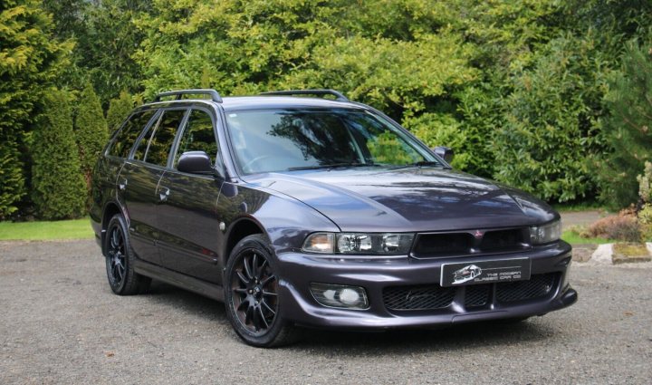 My new (old) 2003 Mitsubishi Galant VI Estate - Page 1 - Readers' Cars - PistonHeads UK - The image showcases a sleek, dark-colored sedan parked on a gravel driveway. The car is a four-door vehicle with a distinctive black roof. It features alloy wheels and a performance exhaust system, hinting at a high-performance or sports model. The setting includes trees in the background and a clear sky above, suggesting an outdoor location during daylight hours.