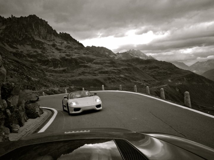 360 thread - Page 1 - Ferrari V8 - PistonHeads - The image captures a serene landscape of rocky terrain and peaks that dominate a distant horizon under a cloudy sky. A curving road lightly dappled with patches of sunshine gracefully through the scene, providing a lovely contrast to the rugged surroundings. A sports car, moving towards us, has just rounded a bend, adding a sense of adventure and urgency to the otherwise peaceful setting. A solitary wall or fence, devoid of foliage, remains steadfast in the middle of the road, perhaps part of a guard rail, ensuring safety for travelers.