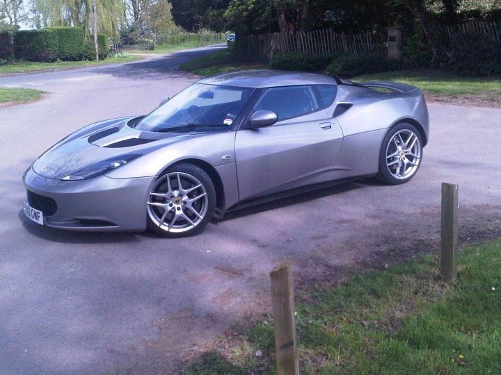 Finally got one - Page 1 - Evora - PistonHeads - The image features a dark gray sports car parked on an asphalt surface. The car appears to be a Lamborghini, characterized by its distinctive sloping front and large air intakes on either side. It has black rims and a license plate on the back, which is clearly visible. The background shows a suburban road with a line of trees and a fence, indicating that the car is parked in a private or residential area. The setting gives the impression of calmness and offers a clear view of the car.