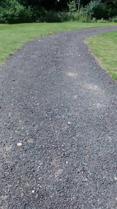 Cheapest way to make a driveway - Page 1 - Homes, Gardens and DIY - PistonHeads - The image captures a serene outdoor scene, featuring a gravel path that winds its way through a lush green field. The path, composed of small, irregularly shaped gravel stones, provides a stark contrast to the surrounding greenery. Encircling the path is a band of grass, unkempt but still verdant. Beyond the path and grass, a field stretches out, its surface carpeted in a thick layer of grass. Trees can be seen in the distance, adding depth and dimension to the scene. The perspective of the photo suggests it was taken standing on the grass. The relative positions of the elements in the image create a sense of depth and perspective.