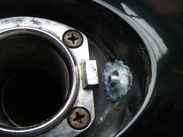 Fuel Filler Cap Security Idea - Page 1 - S Series - PistonHeads - The image shows a close-up of a mechanical part, which appears to be an engine component. There are two visible bolts of the component holding the metal piece together. A threaded post links the metal component to the black plastic backing, which has a star-like texture. A small, rounded cap or grommet is a distinguishing feature on the metal piece. The surface of the component exhibits signs of dust and dirt, indicating it is a used part or has been exposed to elements. There is no text visible in the image.