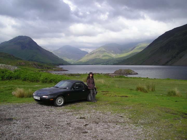 One NA, two holidays, 3000 miles - one change of  heart... - Page 1 - Mazda MX5/Eunos/Miata - PistonHeads - In the image, a woman is standing amidst a field of grass, next to a picturesque lake. The lake is calm and reflective, with a majestic backdrop of rolling hills and distant mountains. A black car is parked on the grass next to her, adding a modern touch to the otherwise tranquil natural setting. The sky above the field is overcast, creating a serene and atmospheric mood in the scene.