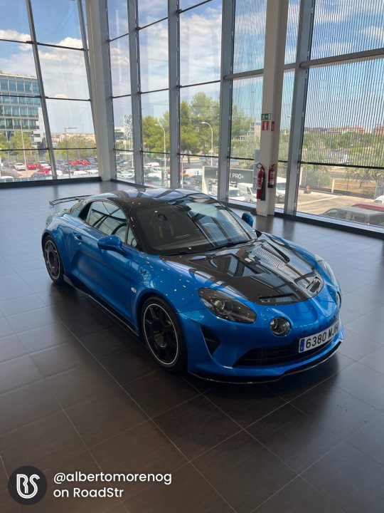 Introducing my new 2023 Alpine A110R  - Page 1 - Readers' Cars - PistonHeads UK - The image shows a room with a large window, through which the outside is visible. A blue sports car is parked inside the room on a black and white striped floor, facing towards the right side of the image. The vehicle features a sleek design with a prominent front grille. There is a person standing in the background, but they are not the focus of the photo. The overall impression is that of an automotive showroom or dealership, where cars are displayed for potential buyers.