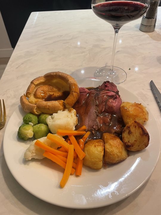 Photo of your dinner (Vol 3) - Page 597 - Food, Drink & Restaurants - PistonHeads UK - The image shows a dining table with a meal laid out. On the table, there is a white plate holding a portion of food consisting of roast beef and various vegetables including carrots and broccoli. Behind the plate, to the left, there's a glass of red wine. In the background, to the right, there is a view of a kitchen with a countertop visible, but no other objects or details can be discerned from this perspective. The overall impression is that of a well-presented meal, suggesting a home setting and possibly a special occasion given the presence of wine.