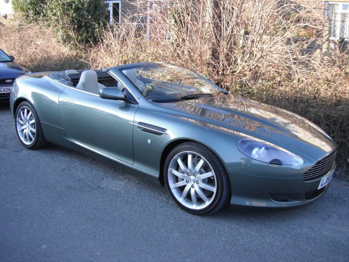 Your DB7 pictures. - Page 1 - Aston Martin - PistonHeads - The image shows a silver convertible car parked on the side of a road. The car's top is down, revealing a blue interior, and a person can be seen seated in the driver's seat. The vehicle is positioned somewhat diagonally to the camera, allowing a view of both its front and its passenger side. The car appears to be in good condition, with shiny chrome accents, sleek lines, and multi-spoke wheels. The surroundings suggest an urban area with buildings and greenery in the backdrop. There are no visible license plates on the car.