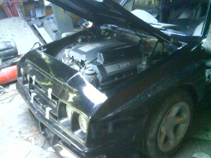 Pistonheads - The image shows the front of a BMW car with the hood open, revealing the engine and other parts. The car is characterized by the round headlights and prominent BMW logo. The car appears to be in a workshop or garage setting, as suggested by the metal workbench in the background. The overall condition of the car suggests it has been used and possibly modified or repaired. There are no texts or distinctive branding visible in the image.