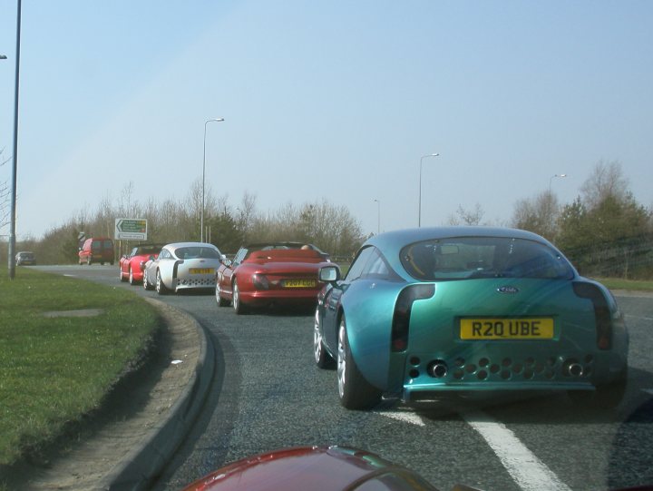 Tvrcc Pistonheads Cobweb Valley Tees - The image shows a busy multi-lane highway with a section of lush green grass visible on the left side of the frame. The primary focus of the photograph is on numerous cars in motion, with a bright blue car prominently featured towards the front. The vehicles are traveling in the same direction, and the scene conveys a sense of rush hour traffic. It appears to be an overcast day with no strong sunlight.