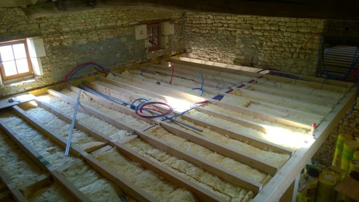 Our French farmhouse build thread. - Page 5 - Homes, Gardens and DIY - PistonHeads - The image shows a construction site with an unfinished floor that resembles a wooden or plywood subfloor. The flooring material is in aome sort of insulation or pre-prep state. The work spans a large area and is framed by wooden side walls which look like part of a construction framework or scaffolding. Wires and tubing are visible, suggesting that utilities and electrical wiring might be in the process of being installed or have been installed in a temporary state. The environment indicates an ongoing construction or renovation project.