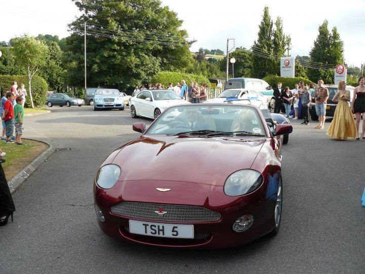 Your DB7 pictures. - Page 1 - Aston Martin - PistonHeads - The image shows a street scene where a red sports car with a license plate reading "TSH 5" is the center of attention. The car is driving down the street and is the focus of a crowd of people standing on the sidewalk. Among the spectators, a woman in a yellow dress is noticeable. The street is busy with other cars, some of which are parked, and it appears to be a vibrant and populous area. The aura of excitement in the eyes of the onlookers implies that this could be a special event or a parade of sorts.