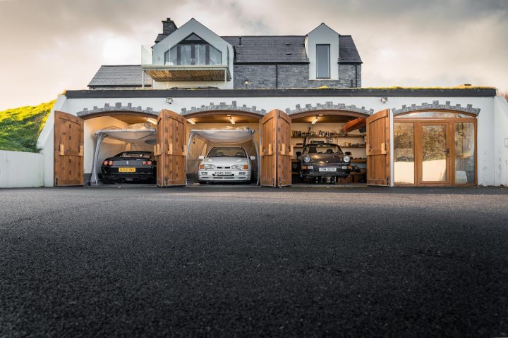 Underground garage build - Page 7 - Homes, Gardens and DIY - PistonHeads UK - The image shows a modern, two-story residential building with a car lift. On the ground floor, there are four cars parked in front of the garage door; on the upper level, there are additional spaces visible through the windows. The exterior of the house has a contemporary design with a combination of stone and wood elements, including a large overhang supported by wooden beams. The sky is overcast, suggesting it might be raining or very cloudy. The car lift is prominently positioned in front of the building, indicating that it may be a feature of the property.