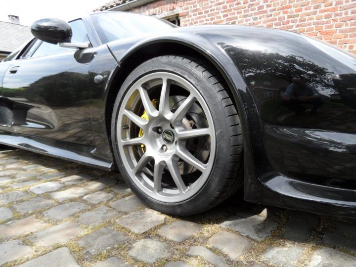 Pistonheads - The image features the front wheel of a black sports car parked on a stone driveway. The car's rims are visible, and it appears to be a luxury vehicle. The stone driveway provides a contrasting texture to the smooth, gleaming car. There are no people or other objects in the immediate vicinity. The car is facing us, the viewers, with the front wheel clearly visible. The overall scene suggests a quiet, upscale setting.