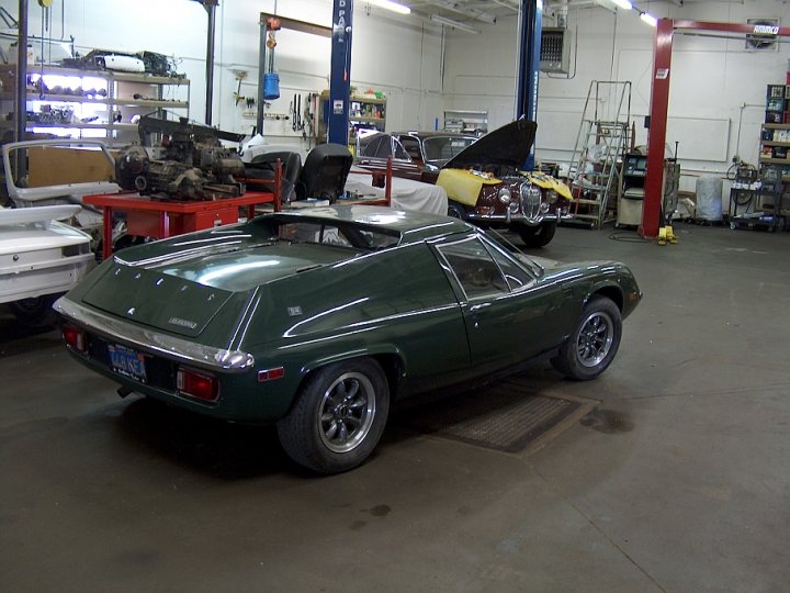 Lotus Europa 1966-1975 & tuning & conversions etc - Page 1 - General Lotus Stuff - PistonHeads - The image shows a spacious garage or workshop filled with various vehicles. The main focus is a green and black sports car with its hood open, revealing its engine. The car is parked close to a red storage rack, and in the background, other cars and machinery can be seen. The lighting in the room is bright, suggesting a professional workshop environment.