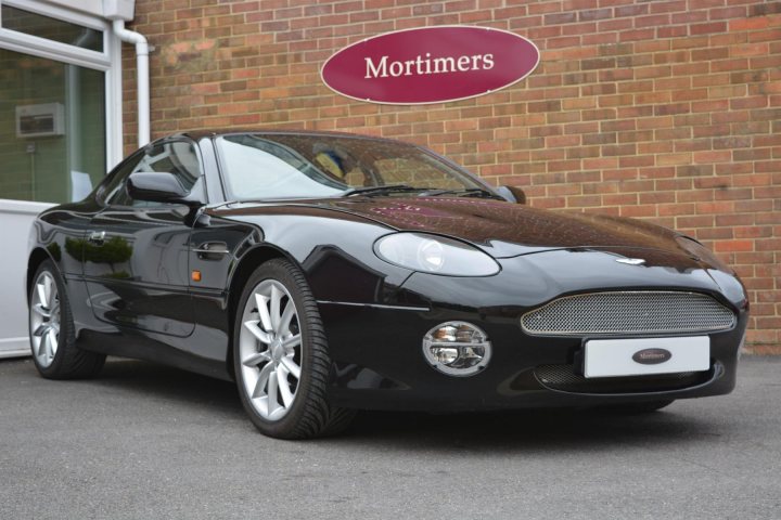 Aston Martin DB7 Vantage - Page 1 - Readers' Cars - PistonHeads UK - The image shows a black convertible sports car parked outdoors. It's positioned in front of a building with a sign that reads "Mortimers," indicating it might be a business or residential establishment. The car is viewed from an angle, displaying the side profile of the vehicle. The car features alloy wheels and has the top down, revealing its interior. There is also an Aston Martin badge visible on the grille of the car, confirming the brand.