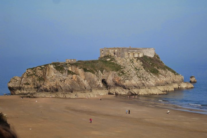 A person on a beach with a kite - Pistonheads - The image captures a breathtaking coastal scene where a massive rock formation juts out from the shoreline into the ocean. Atop this rock, an impressive fortification or castle ruin stands, its stone structure eroded by the elements but still imposing. The sandy beach stretches out to the left, revealing more of the ocean's surface, while the clear, blue sky above suggests a sunny day. In the distance, several small figures appear, likely beachgoers enjoying the natural beauty.