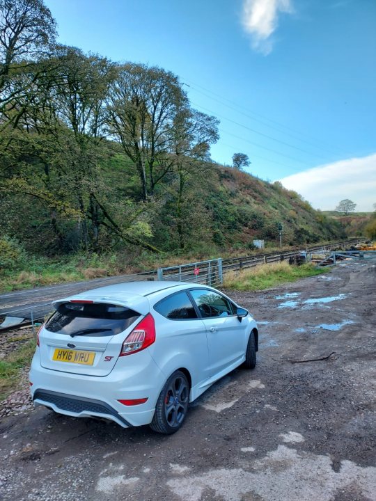 2016 Ford Fiesta ST2  - Page 1 - Readers' Cars - PistonHeads UK - The image shows a white Ford Fiesta parked in an open lot. It has black roof racks and a black rear spoiler. The car is facing the camera, but the license plate is not visible. Behind the car, there is a road that seems to be closed off or unoccupied at this moment. To the left of the frame, there is a grassy area with trees, and to the right, there appears to be some sort of construction site or debris. The sky is overcast, suggesting it might have rained recently. The setting suggests an isolated location or perhaps a place where vehicles are not regularly parked.