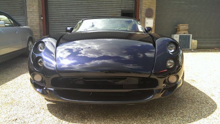 Headlight and rear light conversion - Page 1 - Cerbera - PistonHeads UK - The image shows a sleek, dark-colored sports car parked in front of a building. The vehicle's design suggests it is a convertible with a retractable roof. It has a prominent grille and distinctive headlights. In the background, there are cars and a person visible, indicating that this photo was taken in a parking lot or similar location.