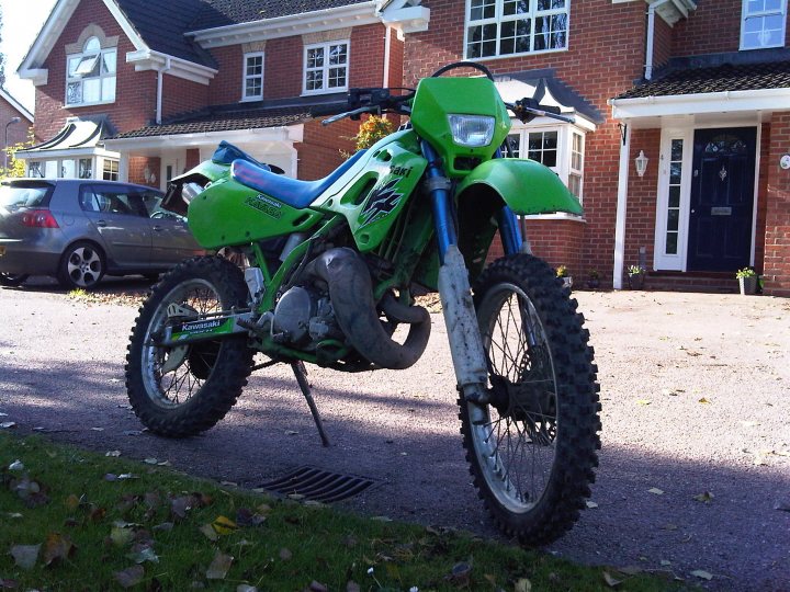 Pistonheads Tidy Kdx Modernisation - This image shows a green dirt bike parked on a concrete driveway. The motorbike has a prominent front wheel, mudguards, and a raised seat. It features a camouflage pattern, indicating it is designed for off-road use. The motorbike is positioned diagonally across the driveway, leaning slightly towards the left side of the frame. Behind the bike, there's a two-story brick house with a porch, a garage with a mostly obscured car, and private driveways of other houses, providing a residential, suburban setting.