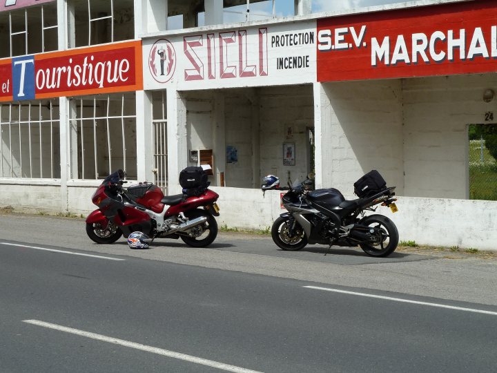 French trip - Page 1 - Aston Martin - PistonHeads - This image presents a quiet street scene. Two motorcycles, a red one with a matching helmet and a second with yellow lighting, are parked side by side on the right side of the frame. Behind them, a white building stands with its red protective signage visible. To the left, a clear blue sky stretches out, suggesting pleasant weather. A sign on the building reads "Touristique" and "Protection Sev.Incendie," indicating its connection to tourism and fire protection services.