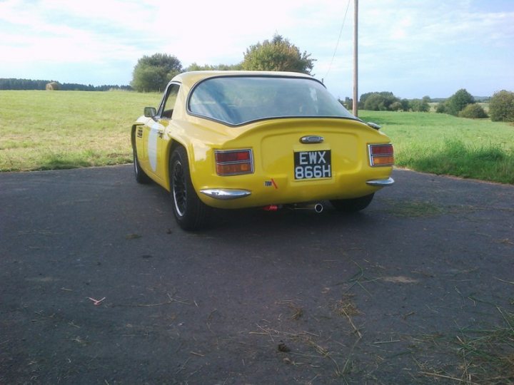 Early TVR Pictures - Page 35 - Classics - PistonHeads - The image shows a classic yellow car, specifically a model that starts with "EXX", parked on a solid, black surface. The car features a distinctive difference in its paint, with the rear end being yellow compared to the distinctive white hood. The rear lights of the car are heritage-style red signal lights, and the side includes UK-style registration plates with "EXX 866H". In the background, the setting is a green field, reinforcing the image's rural atmosphere. There is also a telephone pole visible, suggesting this location is on the outskirts of a populated area.