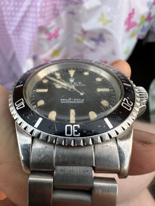 Help with old submariner - Page 1 - Watches - PistonHeads UK - The image captures a scene with an intriguing subject. A hand is seen holding up a silver Rolex watch, which stands out prominently against the rest of the frame. The watch features a black dial and is marked with Roman numerals at 12, 3, 6, and 9 o'clock positions. The silver metal band encircles the watch, adding to its luxurious appearance. The person holding the watch appears to be seated at a table, although only a portion of the table is visible in the image. The backdrop is indistinct, keeping the focus on the Rolex watch held by the individual.