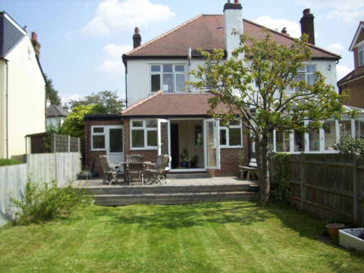 Pistonheads - The image showcases a two-story brick house with a white door and a portico supported by half columns. In the foreground, a well-maintained lawn extends to a wooden deck area, where there's outdoor patio furniture, including chairs and tables, ready for relaxation or entertainment in favorable weather conditions. A tree stands next to the deck, and a section of wooden fence borders the lawn. The sky is hazy, suggesting a soft, possibly overcast weather condition.