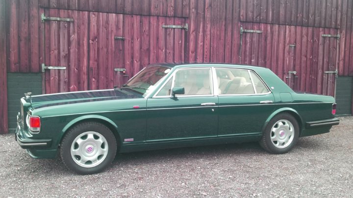 A classic car is parked in a parking lot - Pistonheads - The image showcases a vintage dark blue sedan parked in front of a wooden building with a green roof. The car has a black interior and a manual transmission. The vinyl top is black, matching the color of the front beam bumper and the original carpets. The vehicle is equipped with a four-wheel automatic braking system and power disc brakes. An emblem on the side of the car displays its brand. The car's condition suggests it is well-maintained, with only a few dings and scratches here and there.