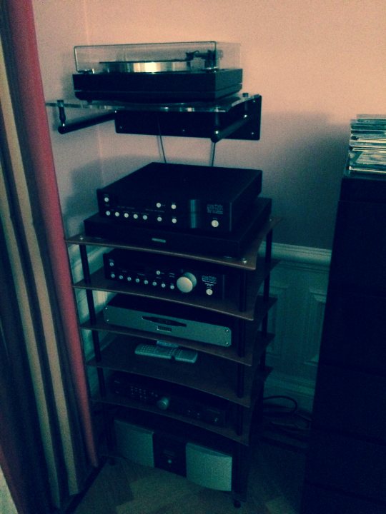 HIFI How old is yours? - Page 3 - Home Cinema & Hi-Fi - PistonHeads - The image shows a collection of audio equipment on a stacked shelf. The turntables at the top appear to be vintage or retro models, suggesting a preference for vinyl sound. Below them, there is a mix of speakers, amplifiers, and other audio components, possibly belonging to an older stereo system. The stack is impeccably organized with each component secured to its designated shelf, reflecting a good deal of personal pride in the setup. The room's pink wall forms a vibrant backdrop to the off-white shelves, creating a contrast in the color scheme.