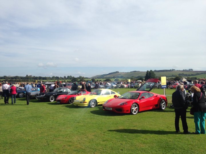 Pistonheads - The image captures a vibrant outdoor scene where a large gathering of people are standing by a row of parked cars. The cars are diverse, including a striking red convertible and several other models, painted in various colors like yellow and white. The people, dressed in casual clothes and carrying handbags, seem to be engaged in conversation or admiring the displayed vehicles. They are standing on a verdant grassy area, with a clear sky overhead. The atmosphere appears to be festive, possibly a car show or a similar event.