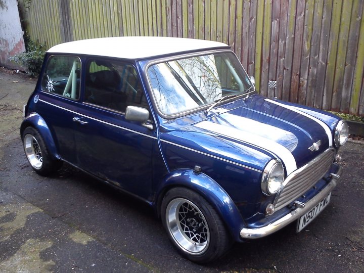 Classic mini - Page 1 - Readers' Cars - PistonHeads - The image features a vintage blue Mini Cooper hard top, parked on a pavement next to a wooden fence. The vehicle is characterized by its distinctive white roof, as well as a silver stripe running down the hood. Its design includes round chrome hubcaps, and there's a white emblem on the front grille. Frosted with antifreeze, the car exhibits a glossy and well-maintained finish.