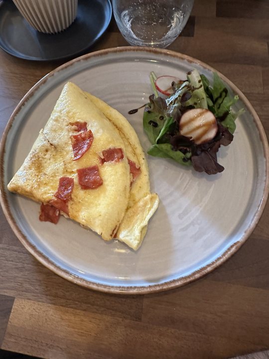 The Great Breakfast photo thread (Vol. 2) - Page 694 - Food, Drink & Restaurants - PistonHeads UK - The image shows a meal served on a plate placed on a table. The plate contains an omelette topped with red peppers and what appears to be bacon, garnished with fresh herbs. Beside the omelette is a small salad with lettuce leaves. The dish is presented on a wooden table with a rustic aesthetic, and there's a white cup visible in the background. The meal looks appetizing and well-prepared, suggesting it could be from a restaurant or a home-cooked meal.