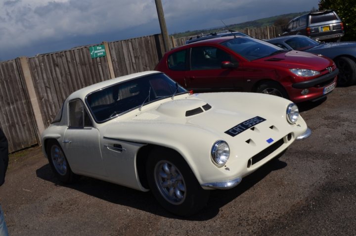 Early TVR Pictures - Page 73 - Classics - PistonHeads - The image shows a scene in a parking lot with a distinctive white racing car with a rounded front, reminiscent of mid-20th century designs. To the right of the racing car, there's a vibrant red car, and on the left side of the frame, we can see part of a person, their figure cut off from the image. The sky overhead is overcast, suggesting a cool or cloudy day. The ground in the parking lot appears to be asphalt.